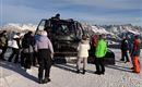 Eine Gruppe von Menschen steht um einen Pistenbully in den Bergen. Im Hintergrund sind schneebedeckte Gipfel und ein klarer Himmel zu sehen.