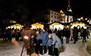 Ein festlicher Weihnachtsmarkt in den Bergen bei Nacht, umgeben von Schnee und beleuchtet von warmen Lichtern. Menschen versammeln sich um Feuerstellen, während im Hintergrund eine Kirche sichtbar ist.