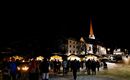 Ein festlicher Weihnachtsmarkt bei Nacht mit vielen Besuchern und stimmungsvollem Licht. Im Hintergrund ist eine Kirche mit einem hohen, spitzen Turm zu sehen.