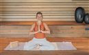 A woman sits in a yoga style on a mat, holding her hands in a prayer position. The room has a wooden appearance and is furnished minimally.