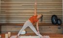 A woman is doing a yoga pose in a bright, modern room. The wooden walls and mats create a relaxing atmosphere.