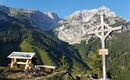 Eine schöne Berglandschaft mit hohen Gipfeln und grünem Wald. Im Vordergrund steht ein Holzkreuz und eine Informationstafel.