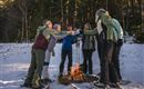 Eine Gruppe von sechs Menschen steht im Schnee um ein Lagerfeuer und stoßt mit Getränken an. Im Hintergrund sind Bäume und eine winterliche Landschaft sichtbar.