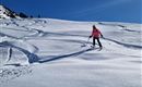 Eine Skifahrerin in einem pinken Outfit fährt über den glitzernden Schnee. Im Hintergrund sind weitere Skifahrer und ein klarer blauer Himmel zu sehen.