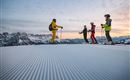Een groep skiërs staat op een vers geprepareerde piste in de bergen. Het uitzicht toont besneeuwde toppen en een heldere hemel bij zonsopgang.