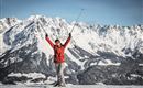 Eine glückliche Skifahrerin steht auf einem schneebedeckten Berg mit den Händen in die Luft. Im Hintergrund sieht man majestätische Berge und eine klare Winterlandschaft.