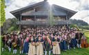 Eine große Gruppe von Menschen in traditioneller Tracht versammelt sich vor einem rustikalen, alpinen Holzhaus. Sie halten verschiedene Instrumente und stehen auf einer grünen Wiese.