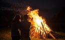 Ein romantisches Lagerfeuer in der Nacht, umgeben von Bäumen. Zwei Personen stehen nebeneinander und beobachten die Flammen.
