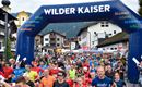 Ein großes Teilnehmerfeld beim Start eines Rennens unter einem blauen Tor mit der Aufschrift "Wilder Kaiser". Viele Läufer in bunten Shirts bewegen sich in Richtung des Starts in einer malerischen Umgebung.