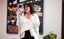 Eine Frau mit Brille singt lächelnd ins Mikrofon. Im Hintergrund sind unscharfe Lichter und eine Pflanze zu sehen.
