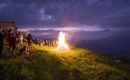 Eine Gruppe von Menschen steht auf einer Wiese und beobachtet ein großes Feuer unter einem bewölkten Himmel. Funken steigen in die Nachtluft auf, während die Szenerie eine festliche Stimmung vermittelt.