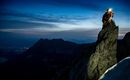 A climber stands on a high rock and inspects his gear. In the background, the mountainous landscapes and a clear sky at dusk are visible.