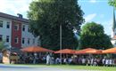 A busy outdoor area with orange umbrellas and many guests eating and drinking together. In the background, a large building and a church tower can be seen.