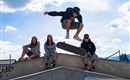 A group of teenagers skateboarding in the skate park. A boy is jumping over the skateboard while the others watch.