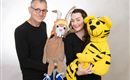 Ein Mann und eine Frau halten jeweils ein Stofftier in den Händen. Der Mann hat eine Puppenfigur und die Frau hält ein gelbes Tiger-Stofftier.