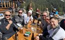 Eine Gruppe von sechs Männern sitzt an einem Tisch im Freien und genießt Getränke. Im Hintergrund sind Bäume und Berge zu sehen, die eine alpine Landschaft darstellen.