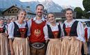 Eine Gruppe von vier Personen in traditioneller Tracht lächelt in die Kamera. Im Hintergrund sind Berge und eine ländliche Umgebung zu sehen.