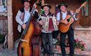 Drei Musiker stehen in traditioneller Tracht und spielen Instrumente. Im Hintergrund ist eine rustikale Holzkonstruktion mit Blumen zu sehen.
