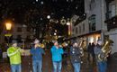 A group of musicians is playing in the snowfall on a festively lit street. The atmosphere is cozy and Christmas-like.