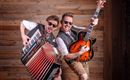 Two men playing music joyfully stand in front of a wooden wall. One plays the accordion while the other holds a guitar, and both are wearing cool sunglasses.
