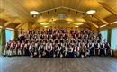Eine große Gruppe von Menschen in traditioneller Tracht steht in einem Holzraum. Sie lächeln und posieren gemeinsam für ein Foto.