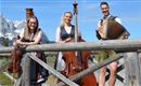 Drei Musiker stehen an einer Holzbrücke in den Bergen. Sie spielen Kontrabass und Akkordeon und tragen traditionelle Trachten.