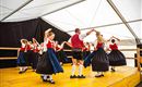 Eine Gruppe von Tänzern in traditionellen Trachten führt einen Volkstanz auf. Sie bewegen sich fröhlich auf einer Bühne in einem Zelt.