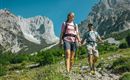Zwei Personen wandern auf einem Weg in den Bergen. Die Landschaft ist grün mit blühenden Blumen und imposanten Felsformationen im Hintergrund.