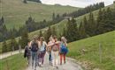 Eine Gruppe von Frauen wandert einen schmalen Weg in einer grünen Berglandschaft. Im Hintergrund sind Bäume und sanfte Hügel zu sehen.
