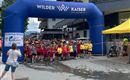 Eine Gruppe von Kindern steht unter einem großen blauen Startbogen, der den Schriftzug "Wilder Kaiser" trägt. Im Hintergrund sind Gebäude und eine gelbe Zeltstation sichtbar.