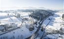 Een schilderachtig winterlandschap met een kasteel te midden van sneeuwbedekte velden. De omgeving is rustig en idillisch, omringd door bergen op de achtergrond.
