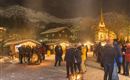 Ein festlicher Weihnachtsmarkt in den Bergen bei Nacht, umgeben von Schnee und beleuchtet von warmen Lichtern. Menschen versammeln sich um Feuerstellen, während im Hintergrund eine Kirche sichtbar ist.
