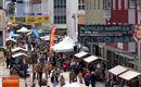 Eine lebhafte Fußgängerzone mit vielen Menschen und Verkaufsständen. Über den Ständen hängen bunte Banner, die auf den Wörgler Markt hinweisen.