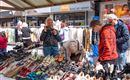 Ein gemütlicher Markt mit einer großen Auswahl an Schuhen und fröhlichen Menschen. Im Hintergrund sind Kleidung und andere Verkaufsstände zu sehen.