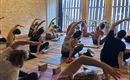 Eine Gruppe von Menschen praktiziert Yoga in einem hellen Raum mit Holzwänden. Sie dehnen sich auf Matten und nutzen Yogablöcke.