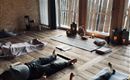 Ein ruhiger Raum mit Holzfußboden, in dem Menschen entspannt auf Yogamatten liegen. Sanftes Licht strömt durch die Fenster und schafft eine entspannende Atmosphäre.