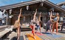 Gruppe von Menschen, die Yoga auf einer Holzterrasse macht. Im Hintergrund ist ein traditionelles Holzhaus zu sehen.