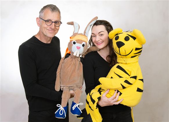 Ein Mann und eine Frau halten jeweils ein Stofftier in den Händen. Der Mann hat eine Puppenfigur und die Frau hält ein gelbes Tiger-Stofftier.