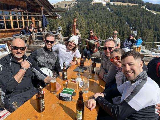 Eine Gruppe von sechs Männern sitzt an einem Tisch im Freien und genießt Getränke. Im Hintergrund sind Bäume und Berge zu sehen, die eine alpine Landschaft darstellen.