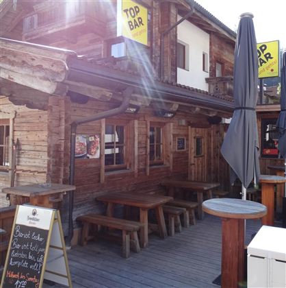 A cozy bar in alpine style with wood paneling and an outdoor terrace. Large signs with the inscription "TOP BAR" hang above the bar.