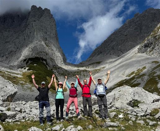 Eine Gruppe von fünf Wanderern steht inmitten einer malerischen Berglandschaft. Sie feiern ihren Erfolg, indem sie die Hände in die Luft heben.