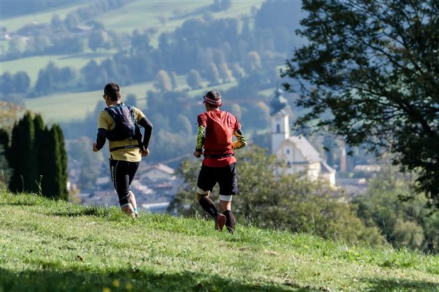 Zwei Läufer joggen über eine grüne Wiese. Im Hintergrund sind sanfte Hügel und ein kleiner Ort mit einer Kirche zu sehen.