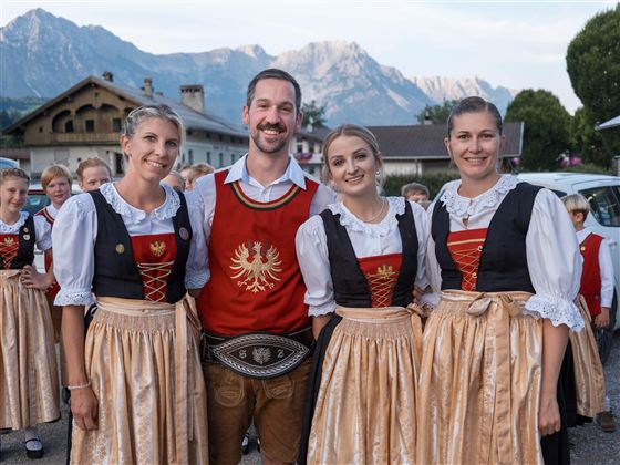 Eine Gruppe von vier Personen in traditioneller Tracht lächelt in die Kamera. Im Hintergrund sind Berge und eine ländliche Umgebung zu sehen.