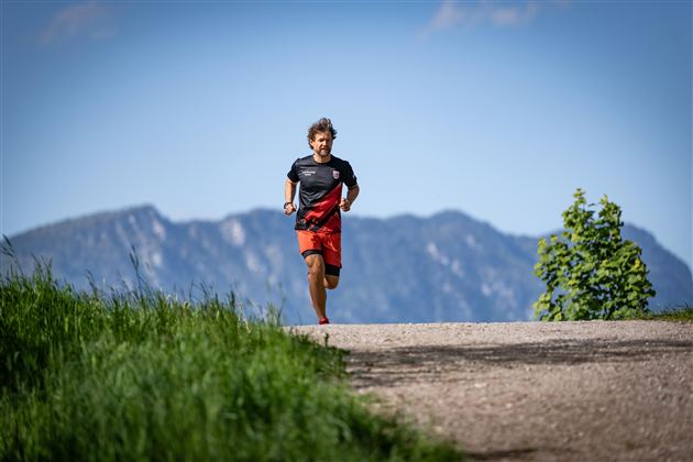 A runner jogs on a path with green vegetation. In the background, mountains and blue sky are visible.