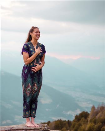 Eine lächelnde Frau steht barfuß auf einem Felsen in der Natur. Sie trägt ein buntes, gemustertes Outfit und blickt in die Ferne.