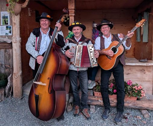 Drei Musiker stehen in traditioneller Tracht und spielen Instrumente. Im Hintergrund ist eine rustikale Holzkonstruktion mit Blumen zu sehen.