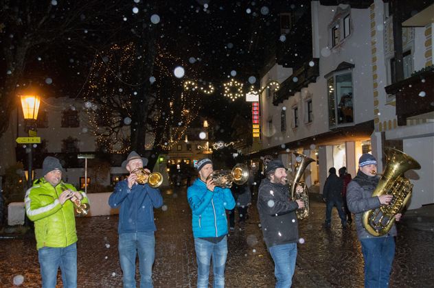 A group of musicians is playing in the snowfall on a festively lit street. The atmosphere is cozy and Christmas-like.