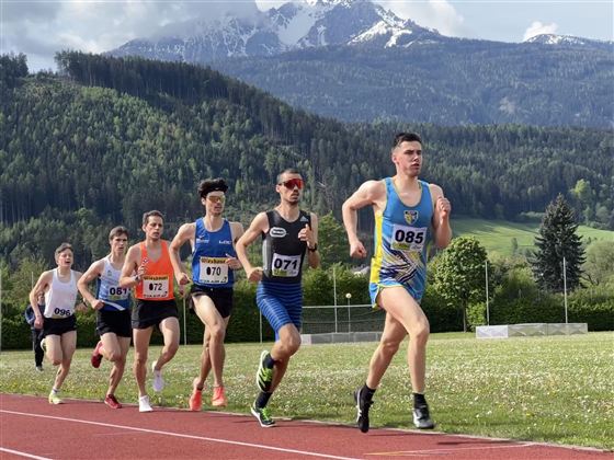 Skupina běžců trénuje na atletické dráze. V pozadí jsou vidět hory a zelené louky.