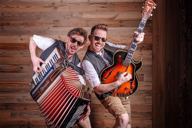 Two musicians happily pose in front of a wooden wall. One plays the accordion and the other holds a guitar.