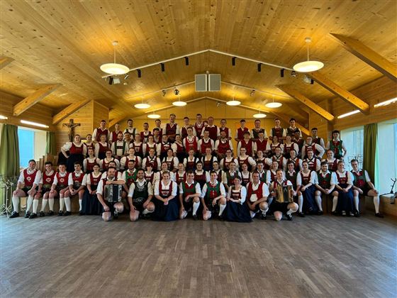 Eine große Gruppe von Menschen in traditioneller Tracht steht in einem Holzraum. Sie lächeln und posieren gemeinsam für ein Foto.
